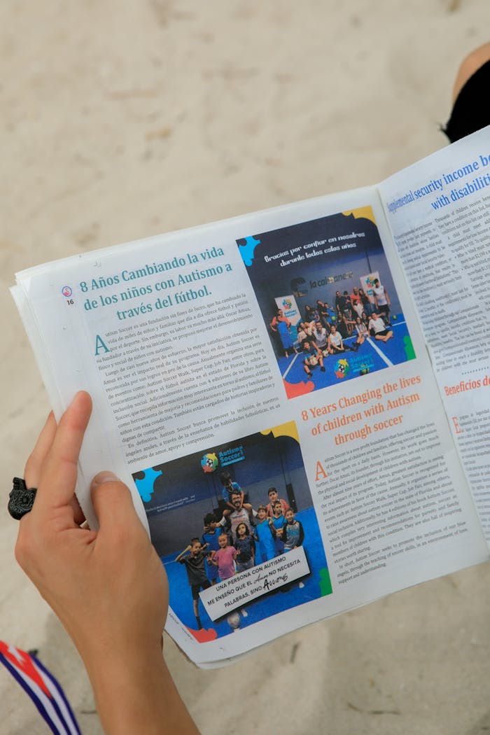 Close-up of hands holding a magazine about autism support through soccer on a sandy beach.